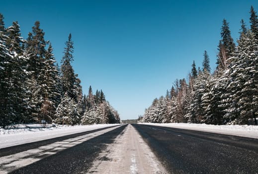 A long stretch of a deserted highway runs through a dense pine forest, blanketed in snow