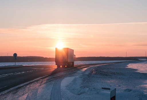 A semi truck cruises down a wintry highway as the sun sets on the horizon, casting a warm glow over the icy road and surrounding landscape.