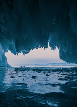 Inside a stunning ice cave on Lake Baikal, large icicles hang from the ceiling, creating a breathtaking winter landscape. Snow-covered mountains can be seen far in the distance.
