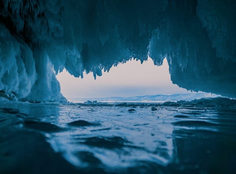 Inside a stunning ice cave on Lake Baikal, large icicles hang from the ceiling, creating a breathtaking winter landscape. Snow-covered mountains can be seen far in the distance.