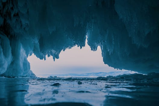 Inside a stunning ice cave on Lake Baikal, large icicles hang from the ceiling, creating a breathtaking winter landscape. Snow-covered mountains can be seen far in the distance.