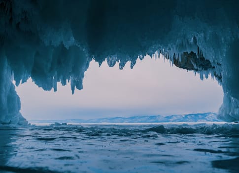 Inside a stunning ice cave on Lake Baikal, large icicles hang from the ceiling, creating a breathtaking winter landscape. Snow-covered mountains can be seen far in the distance.