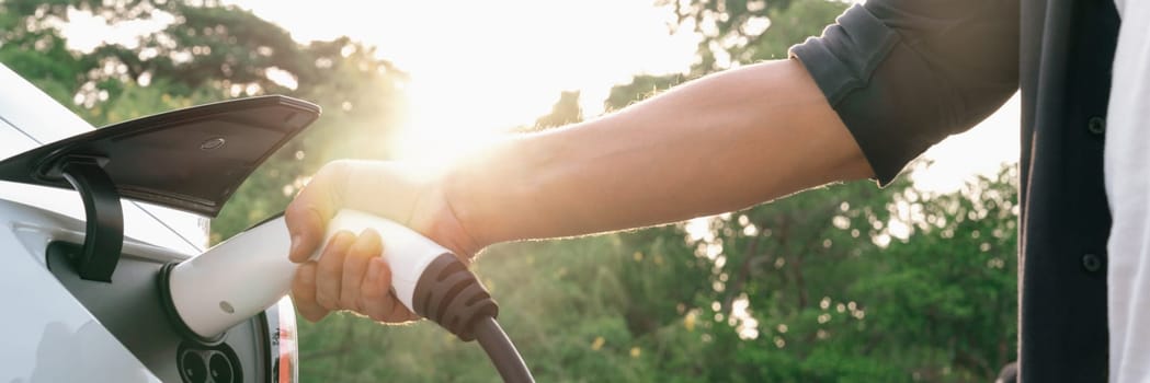 Man recharging battery for electric car during road trip travel EV car in natural forest or national park with sunrise lighting. Eco friendly travel during vacation and holiday. Panorama Exalt