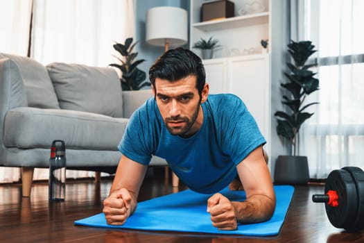 Athletic and sporty man doing plank on fitness mat during home body workout exercise session for fit physique and healthy sport lifestyle at home. Gaiety home exercise workout training.