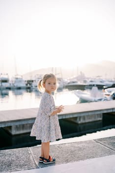 Little girl stands on the pier, moving her hands and looking to the side. High quality photo