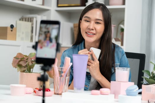 Young beautiful Asian reviewing cosmetic makeup and skin care collection, talking customer on social media online, presenting product surrounded beauty merchandise recording blurred phone . Stratagem.