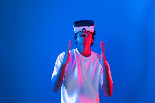 Young African American player wearing VR raising fist up wining game in hologram on blue pink neon lighting wall connecting digital futuristic technology virtual reality metaverse world. Contrivance.