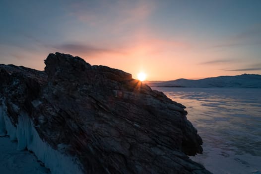 Aerial over the rocky island in lake Baikal. Winter landscape of frozen Baikal at beautiful orange sunrise. Sun reflections on the ice. Popular tourist spot.