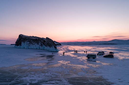 Aerial winter landscape of frozen lake Baikal. Groups of tourists got off the cars and walking around the rocky island in lake Baikal, walking on the crystal clear ice at sunrise. Famous tourist spot