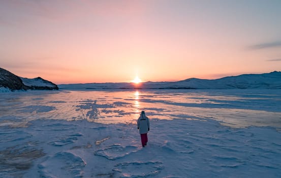 Aerial view on the young woman walking on the blue cracked ice of Baikal at beautiful orange sunrise. Sun reflections on the ice. Winter landscape of frozen Baikal.