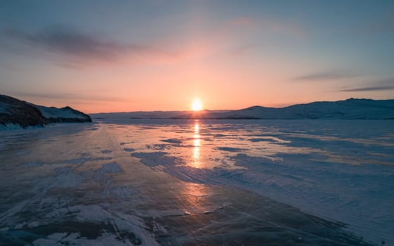 Aerial over the snowy cracky ice of lake Baikal and the rocky island. Winter landscape of frozen Baikal at beautiful lilac sunrise. Sun reflections on the ice. Popular tourist spot. Tilt shot