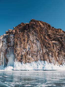 A majestic rocky cliff towers over the frozen surface of Lake Baikal, adorned with stunning ice formations and illuminated under a clear blue sky.