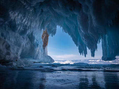 Inside a stunning ice cave on Lake Baikal, large icicles hang from the ceiling, creating a breathtaking winter landscape. Snow-covered mountains can be seen far in the distance.