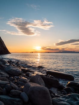 A tranquil sunset casts warm hues over a rocky beach and silhouetted cliffs. The calm ocean waters reflect the changing colors of the sky, creating a serene and picturesque coastal scene.