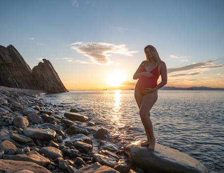 A pregnant woman stands on a rock by the seaside during sunrise, lovingly cradling her baby bump.