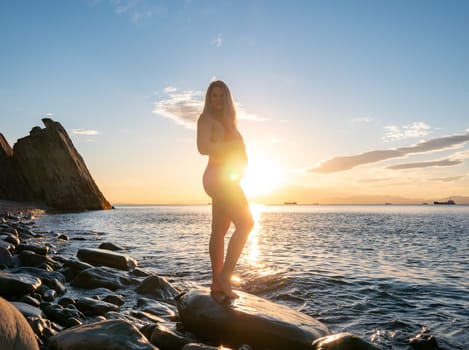 A pregnant woman stands on a rock by the seaside during sunrise, lovingly cradling her baby bump.
