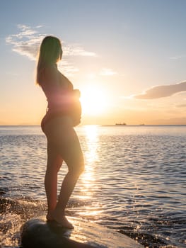 A pregnant woman stands on a rock by the seaside during sunrise, lovingly cradling her baby bump.