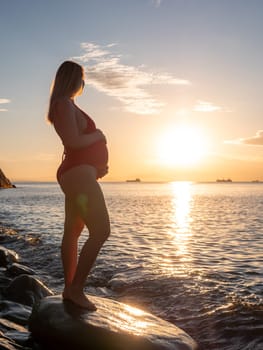 A pregnant woman stands on a rock by the seaside during sunrise, lovingly cradling her baby bump.