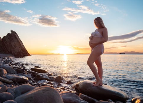 A pregnant woman in a white bikini stands on a rocky beach at sunrise, gently holding her belly. The background features calm water and a distant mountain range.