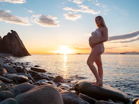 A pregnant woman in a white bikini stands on a rocky beach at sunrise, gently holding her belly. The background features calm water and a distant mountain range.