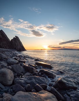 A tranquil sunset casts warm hues over a rocky beach and silhouetted cliffs. The calm ocean waters reflect the changing colors of the sky, creating a serene and picturesque coastal scene.