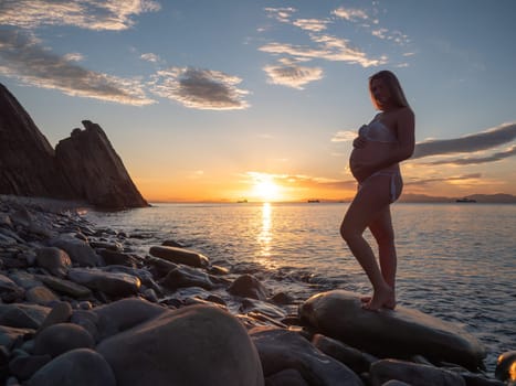 A pregnant woman in a white bikini stands on a rocky beach at sunrise, gently holding her belly. The background features calm water and a distant mountain range.