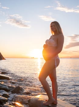 A pregnant woman in a white bikini stands on a rocky beach at sunrise, gently holding her belly. The background features calm water and a distant mountain range.