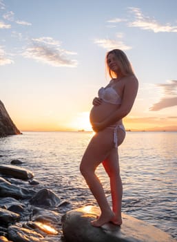 A pregnant woman in a white bikini stands on a rocky beach at sunrise, gently holding her belly. The background features calm water and a distant mountain range.