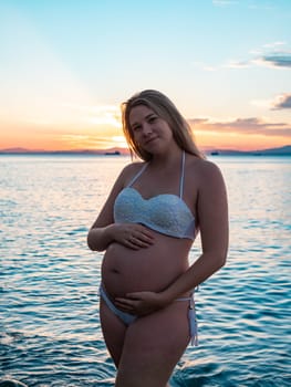 A pregnant woman in a white bikini stands on a rocky beach at sunrise, gently holding her belly. The background features calm water and a distant mountain range.