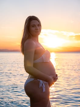 A pregnant woman in a white bikini stands on a rocky beach at sunrise, gently holding her belly. The background features calm water and a distant mountain range.