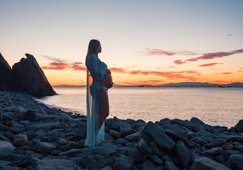 A pregnant woman in a white bikini stands on a rocky beach at sunrise, gently holding her belly. The background features calm water and a distant mountain range.