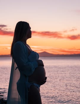 A pregnant woman in a white bikini stands on a rocky beach at sunrise, gently holding her belly. The background features calm water and a distant mountain range.