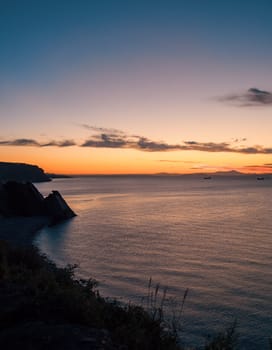 A tranquil sunset casts warm hues over a rocky beach and silhouetted cliffs. The calm ocean waters reflect the changing colors of the sky, creating a serene and picturesque coastal scene.