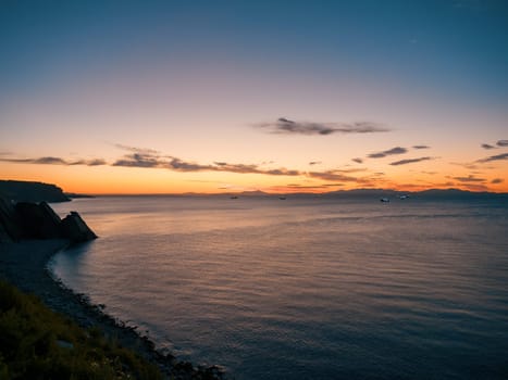 A tranquil sunset casts warm hues over a rocky beach and silhouetted cliffs. The calm ocean waters reflect the changing colors of the sky, creating a serene and picturesque coastal scene.