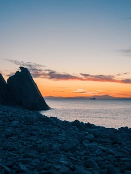 A tranquil sunset casts warm hues over a rocky beach and silhouetted cliffs. The calm ocean waters reflect the changing colors of the sky, creating a serene and picturesque coastal scene.