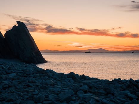 A tranquil sunset casts warm hues over a rocky beach and silhouetted cliffs. The calm ocean waters reflect the changing colors of the sky, creating a serene and picturesque coastal scene.