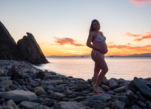 A pregnant woman in a white bikini stands on a rocky beach at sunrise, gently holding her belly. The background features calm water and a distant mountain range.