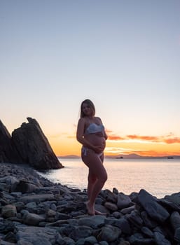 A pregnant woman in a white bikini stands on a rocky beach at sunrise, gently holding her belly. The background features calm water and a distant mountain range.