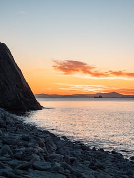 A tranquil sunset casts warm hues over a rocky beach and silhouetted cliffs. The calm ocean waters reflect the changing colors of the sky, creating a serene and picturesque coastal scene.