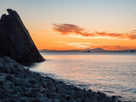 A tranquil sunset casts warm hues over a rocky beach and silhouetted cliffs. The calm ocean waters reflect the changing colors of the sky, creating a serene and picturesque coastal scene.