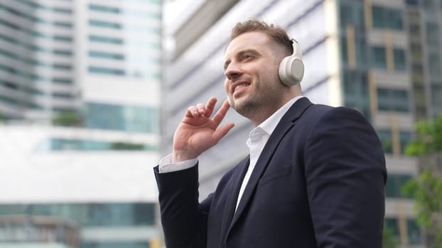 Close up of male leader listen headphone and dancing to relaxing music with cheerful and motivation. Skilled project manager enjoy to listen relaxed song while hold smart phone at urban city. Urbane.