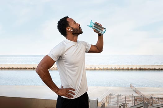 Young Black male runner taking a break to hydrate. African American man drinking water after exercising and jogging outdoors. Healthy lifestyle and sport concept.