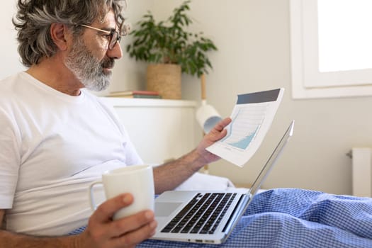 Mature man working with laptop sitting on bed at home. Male drinking coffee and looking at documents. Working from home concept.