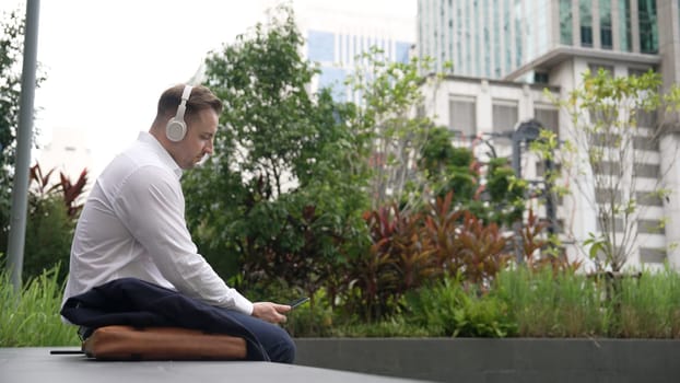 Businessman wear his headphone to listen relaxed music while take off suit and move to music at green city. Manager with headset dancing movement with lively song while hold phone. Back view. Urbane.
