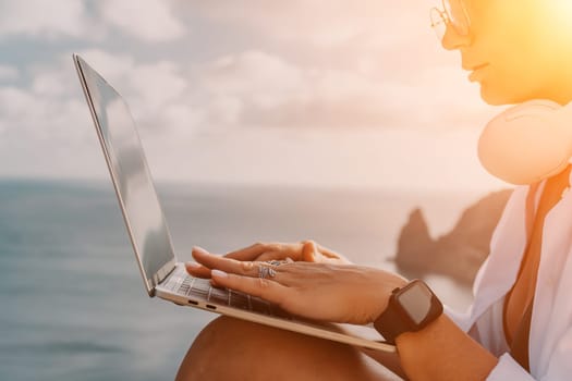 Digital nomad, woman in the hat, a business woman with a laptop sits on the rocks by the sea during sunset, makes a business transaction online from a distance. Freelance, remote work on vacation.