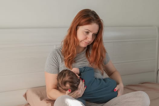 An unhappy woman breastfeeds her son and sits on the bed. Postpartum depression