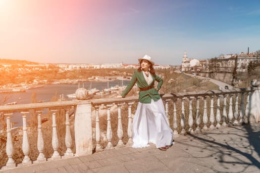 Woman walks around the city, lifestyle. Happy woman in a green jacket, white skirt and hat is sitting on a white fence with balusters overlooking the sea bay and the city