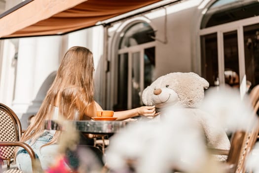 A woman sits cafe with a teddy bear next to her. The scene is set in a city with several chairs and tables around her. The woman is enjoying her time at the outdoor cafe