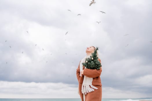 Blond woman Christmas sea. Christmas portrait of a happy woman walking along the beach and holding a Christmas tree on her shoulder. She is wearing a brown coat and a white suit