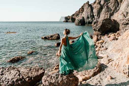 Woman green dress sea. Woman in a long mint dress posing on a beach with rocks on sunny day. Girl on the nature on blue sky background